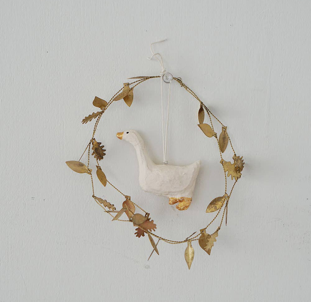 A rustic decorative wreath made of thin brown twine and dried gold-toned leaves, with a small white goose figurine hanging in the center against a plain white wall.