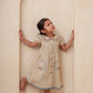 Young girl in a beige dress with floral patterns standing inside an archway.