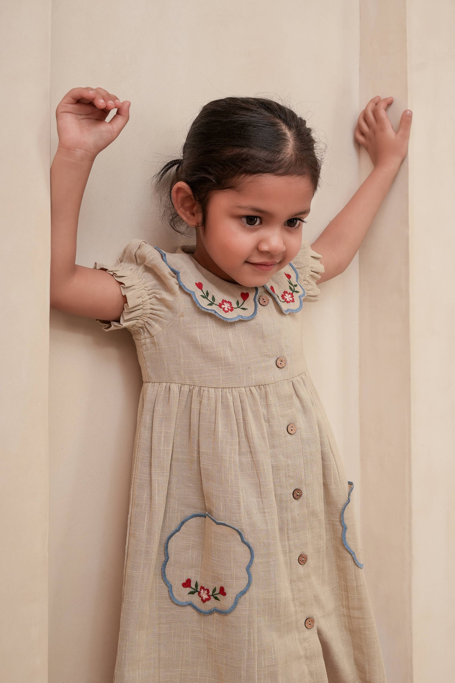 Young girl wearing a beige dress with floral embroidery against a plain background