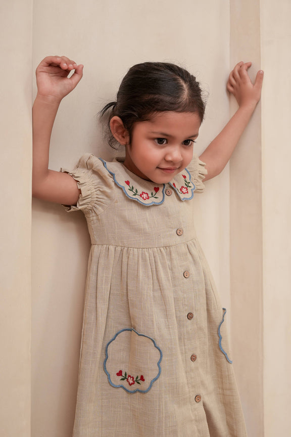 Young girl wearing a beige dress with floral embroidery against a plain background