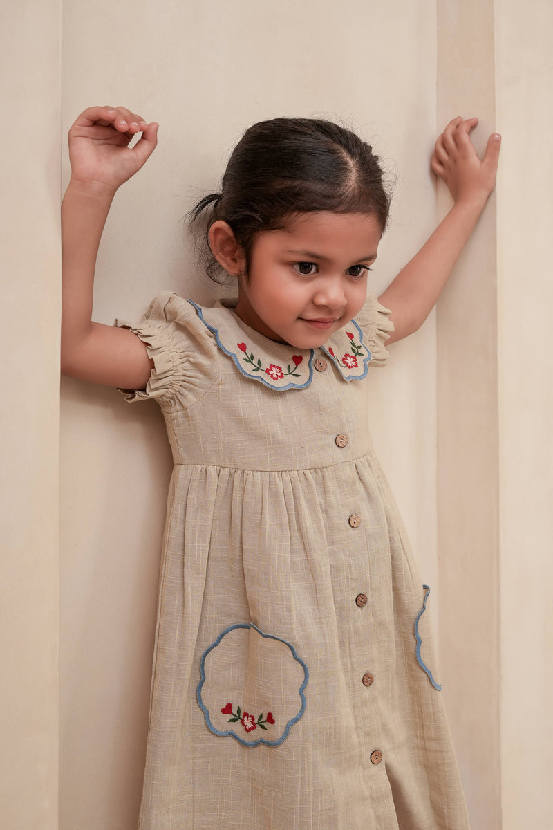 Young girl wearing a beige dress with floral embroidery against a plain background