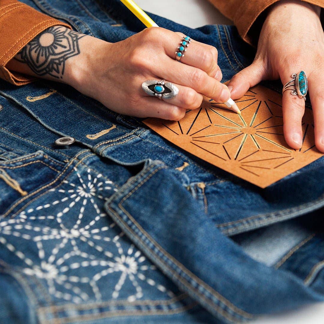Person with tattoos and rings using a pencil on a piece of paper with geometric designs, wearing blue jeans.