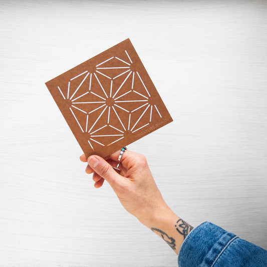 Hand holding a brown square with white geometric pattern against a light gray background