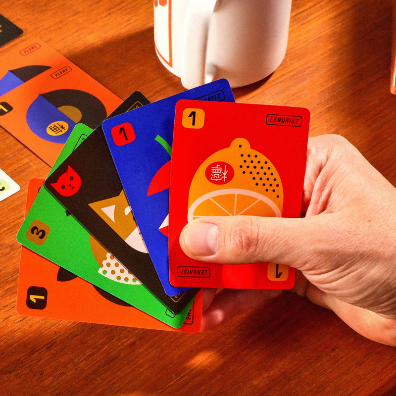 Hand holding colorful playing cards on a wooden table with a white mug in the background.