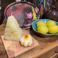 Candle shaped like a cheese wedge with a garlic candle next to in on a wooden surface with decorative plates and a bowl of lemons in the background.
