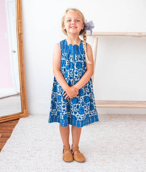 Young girl wearing a blue floral dress standing in a room with a mirror and white wall.