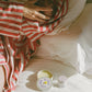 Person holding a baby wrapped in red and white striped blanket with two small containers of balm on a bed.