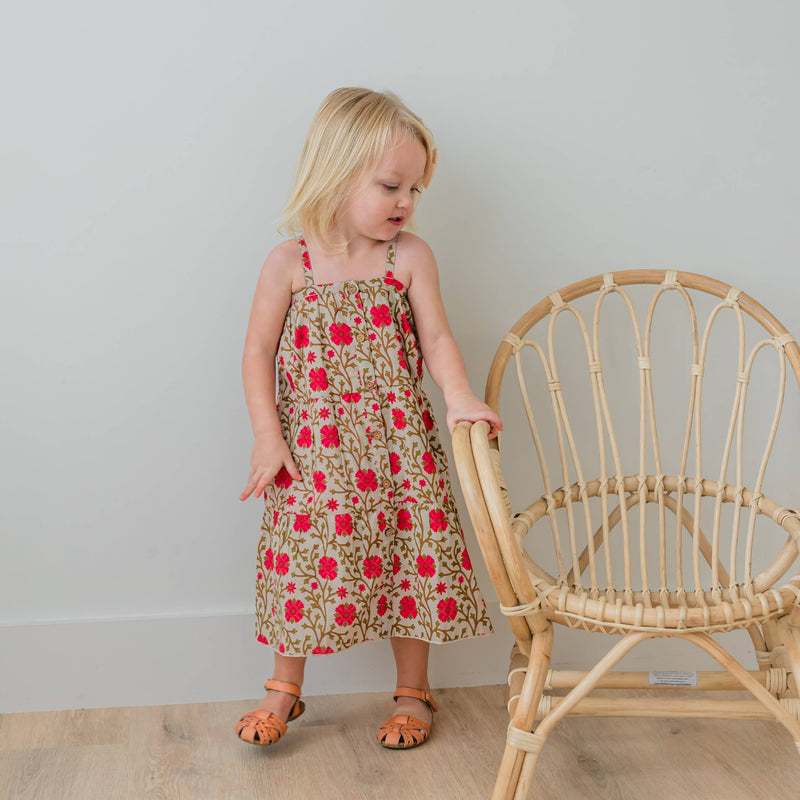 Young girl in a floral dress standing next to a wicker chair against a plain wall.