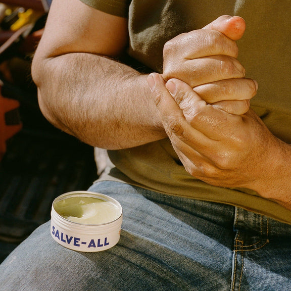 Person applying salve to their hand with a container labeled 'Salve-All' on a blurred background