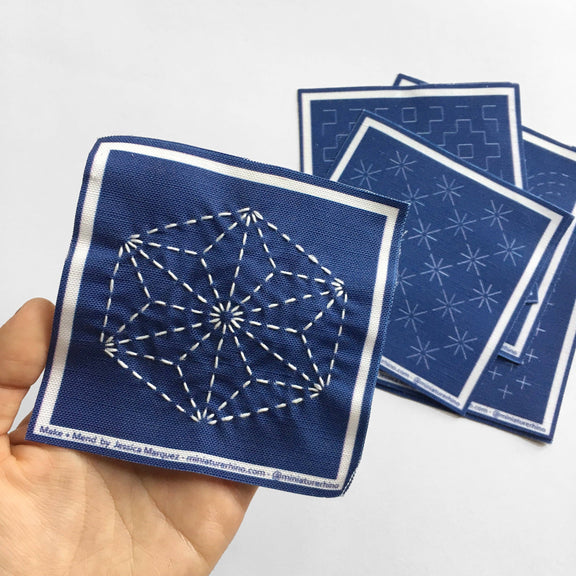 Blue fabric square with white embroidery held by a hand, with additional fabric squares in the background.