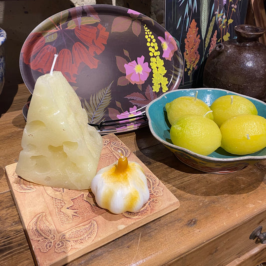 Candle shaped like a a cheese wedge on a wooden surface with decorative plates and a bowl of lemons in the background.
