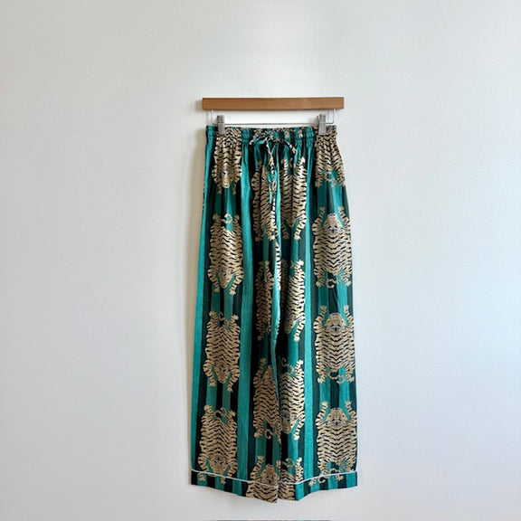 Teal pants with gold patterns hanging on a wooden hanger against a white wall.