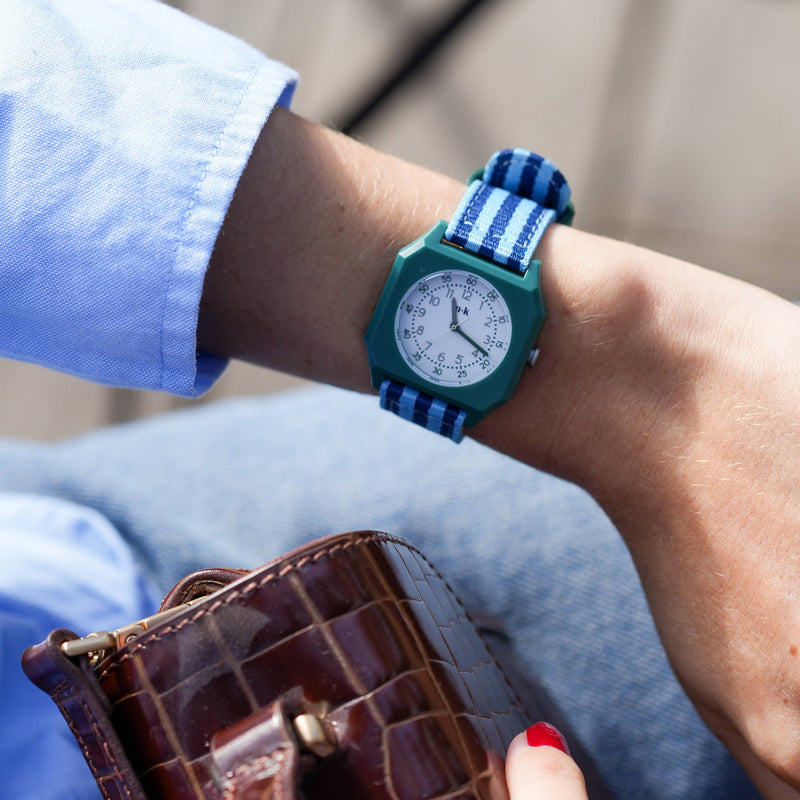Person wearing a teal watch with a blue strap, holding a brown wallet.