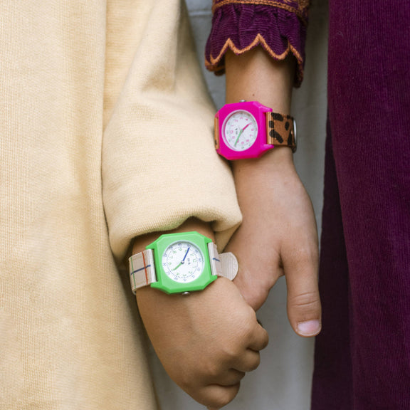 Two hands holding each other, one wearing a green watch and the other a pink watch, against a plain background.