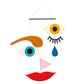 A colorful abstract wall hanging or mobile with stylized facial features: two eyes, one brown eyebrow, a blue teardrop, a pink triangle, and red lips suspended from a black bar against a white background.