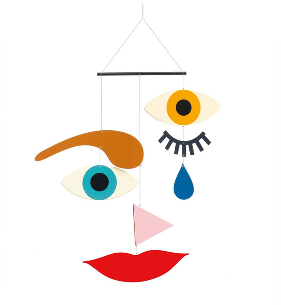A colorful abstract wall hanging or mobile with stylized facial features: two eyes, one brown eyebrow, a blue teardrop, a pink triangle, and red lips suspended from a black bar against a white background.
