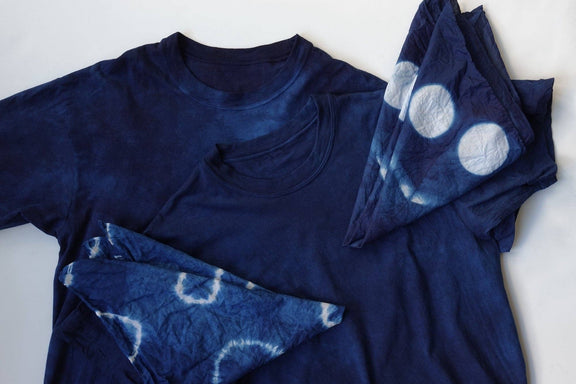 A dark indigo blue tie-dye t-shirt with matching folded fabric pieces showing white tie-dye circle patterns, laid flat on a white background.