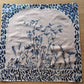 A square fabric napkin or small cloth with a blue floral and leafy design on a light beige background, laid flat on a wooden surface.