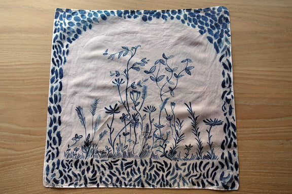 A square fabric napkin or small cloth with a blue floral and leafy design on a light beige background, laid flat on a wooden surface.