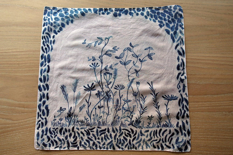 A square fabric napkin or small cloth with a blue floral and leafy design on a light beige background, laid flat on a wooden surface.