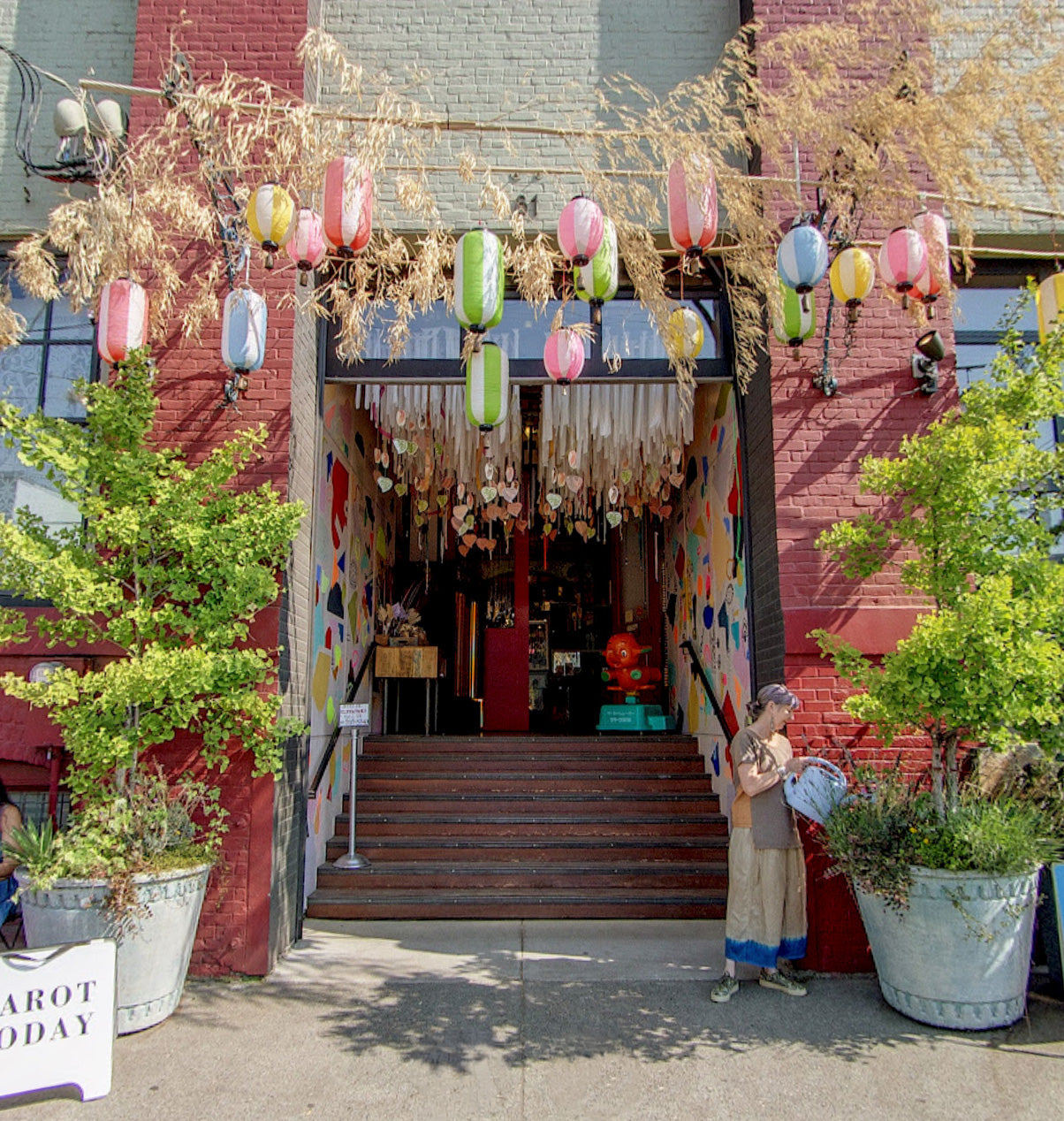 Street front image of a store with a red brick wall and colorful lanterns and a woman watering plants. 