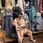 Person in a patterned outfit standing in a clothing store with various garments and accessories.